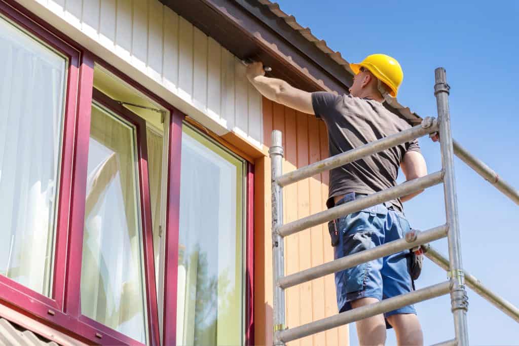 A construction worker wearing a yellow hard hat and denim shorts stands on scaffolding, painting the exterior wall of a house. Specializing in both painting and power washing Kane County IL homes, the scene is completed with large windows framed in red against a clear blue sky.
