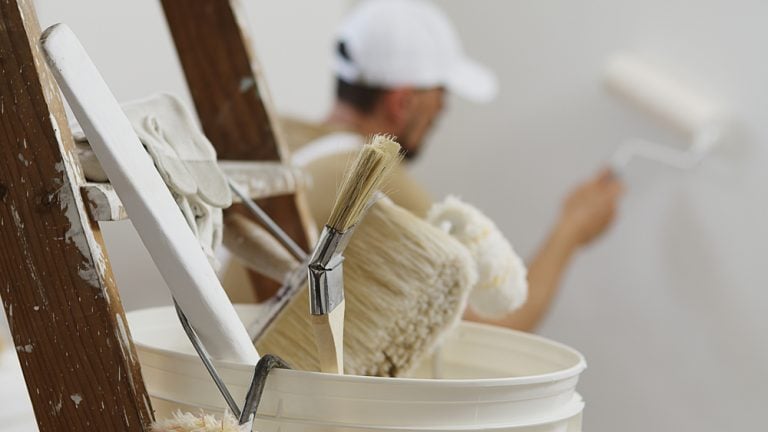 Worker Using Paint Roller On Wall Near A Bucket Of Paint With Brush