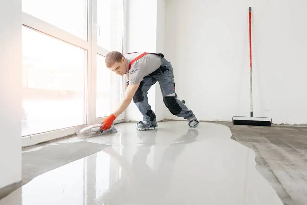 A worker wearing protective gear applies epoxy resin to a floor using a roller, reminiscent of precision found in power washing in Kane County, IL. The empty room is bathed in natural light from large windows, while a broom rests quietly against the wall.