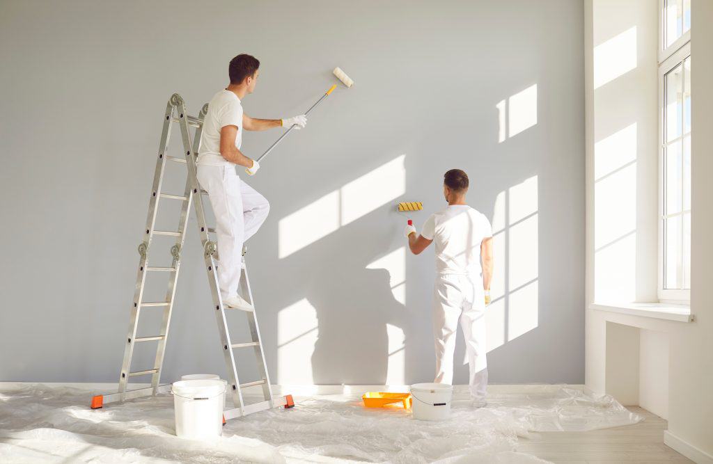 Two people in white clothing paint a wall light gray, embracing their task with care. One stands on a ladder using a roller, while the other holds a small roller near the wall. The floor is covered with plastic, and paint supplies are scattered around as sunlight streams in through large windows—capturing the spirit of painting and power washing Kane County IL.