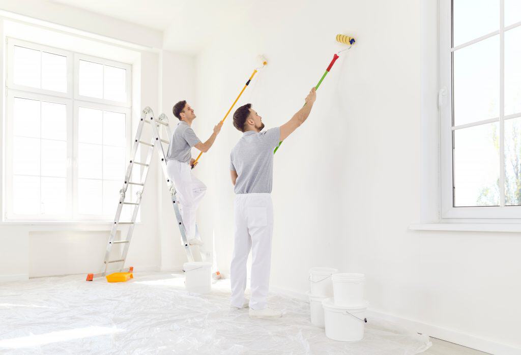 Two people painting a white interior wall. One person stands on a ladder using a roller, while the other paints next to them. The room is bright with large windows, covered floors, and paint buckets around.