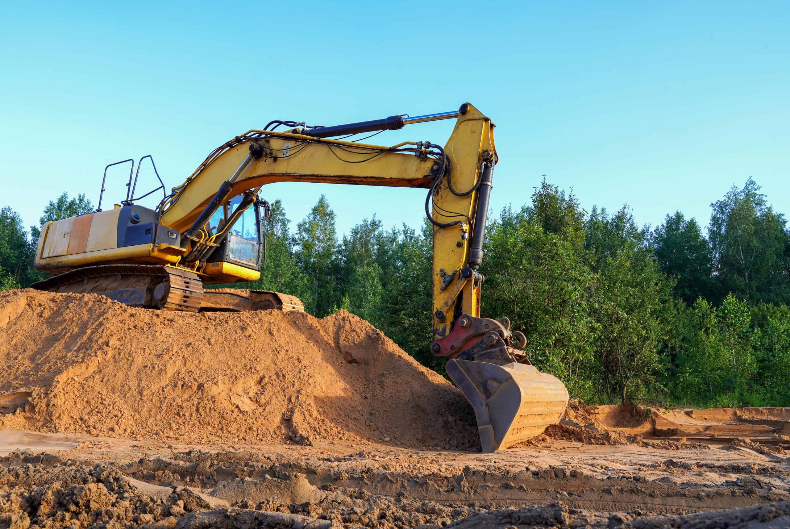 A yellow excavator sits on a pile of sand at a construction site, surrounded by lush green trees under a clear blue sky. The excavator's arm is extended, with its bucket resting on the sand, ready for action like the power washing crews in Kane County, IL.