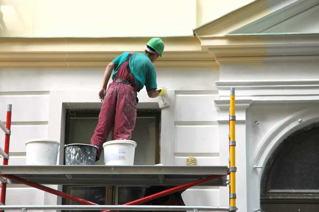 A construction worker wearing a green helmet and red overalls is standing on scaffolding, expertly painting the ornate exterior wall of a building. Nearby, several paint buckets rest on the platform, ready for use in their ongoing painting and power washing project in Kane County, IL.