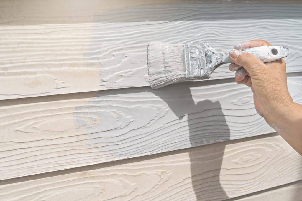 A hand holding a paintbrush applies white paint to the woodgrain-textured siding of a house, freshly prepped after painting and power washing in Kane County, IL. The sun casts shadows over the clean surface, highlighting its renewed look.