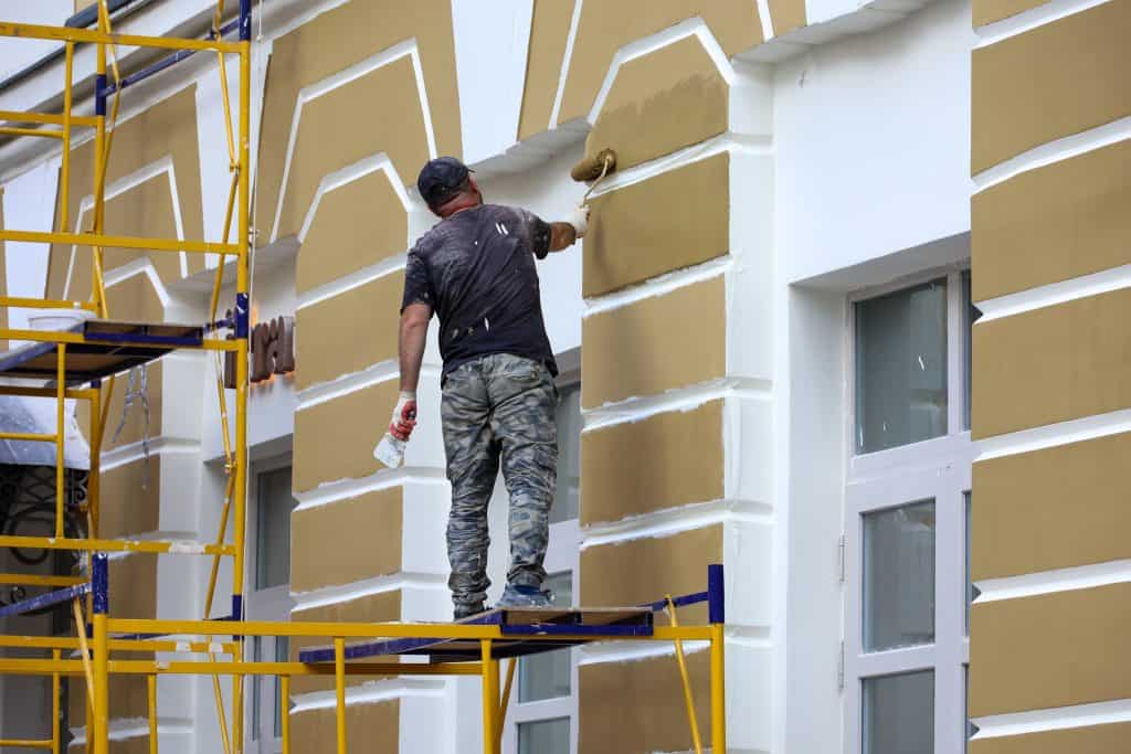 A worker stands on scaffolding, painting a building's facade with gold and white colors. He wears a black cap, dark shirt, and camouflage pants. The building features rectangular patterns around the windows.