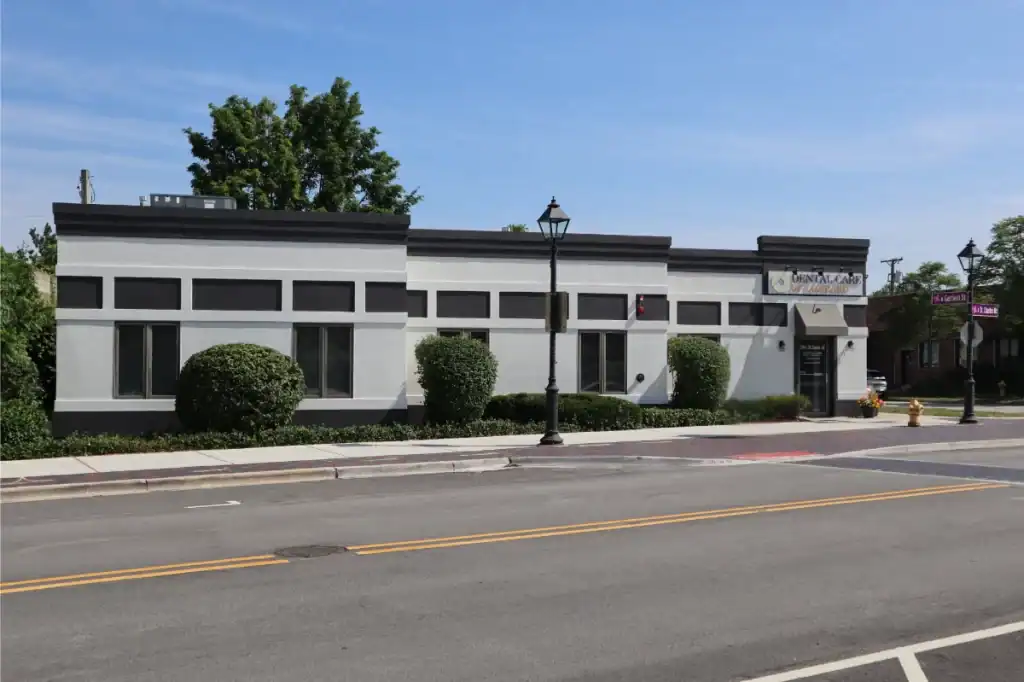 A single-story, white commercial building with dark trim and rectangular windows, landscaped with bushes, on a quiet street with a sidewalk and streetlights under a clear blue sky.