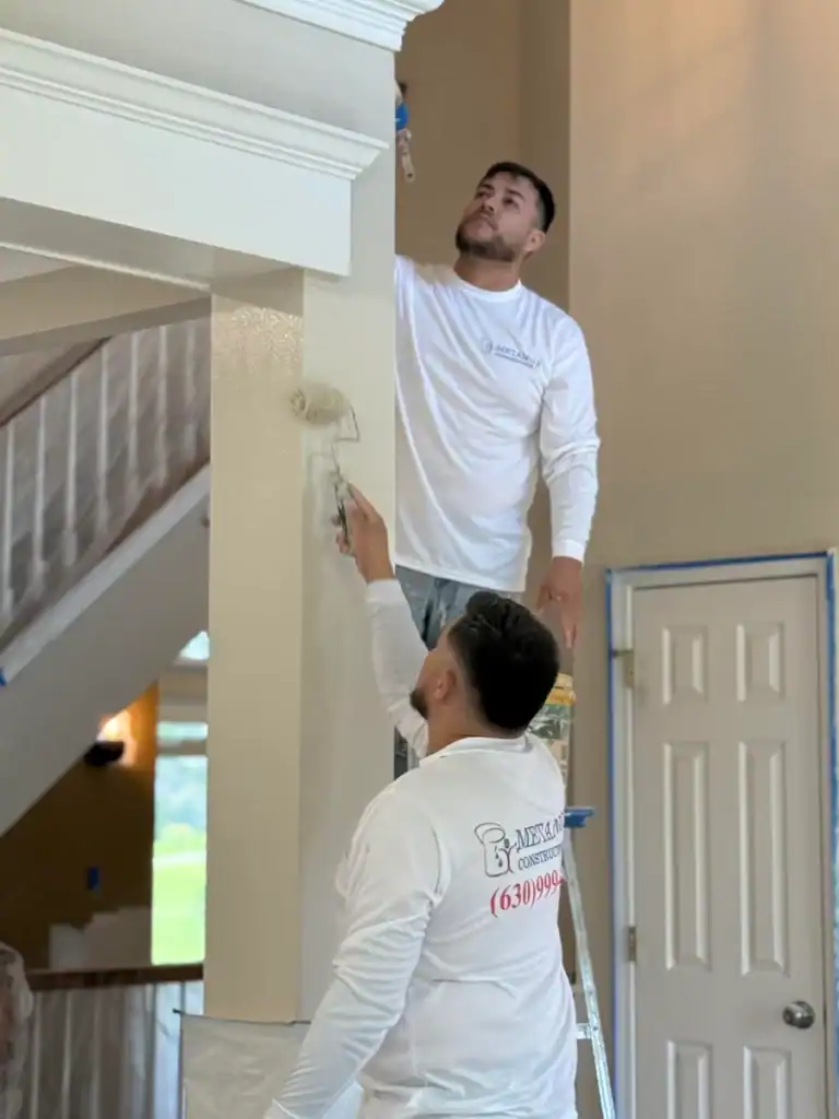 Two men wearing white shirts are painting the walls of a house interior with paint rollers. One man is standing on a ladder painting near the ceiling, while the other paints a wall below him. The area is prepared for painting.