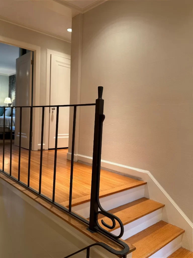 A small staircase with wooden steps and a black metal railing leads up to a hallway with light-colored walls and an open white door. A lamp and part of a bed are visible in an adjoining room.