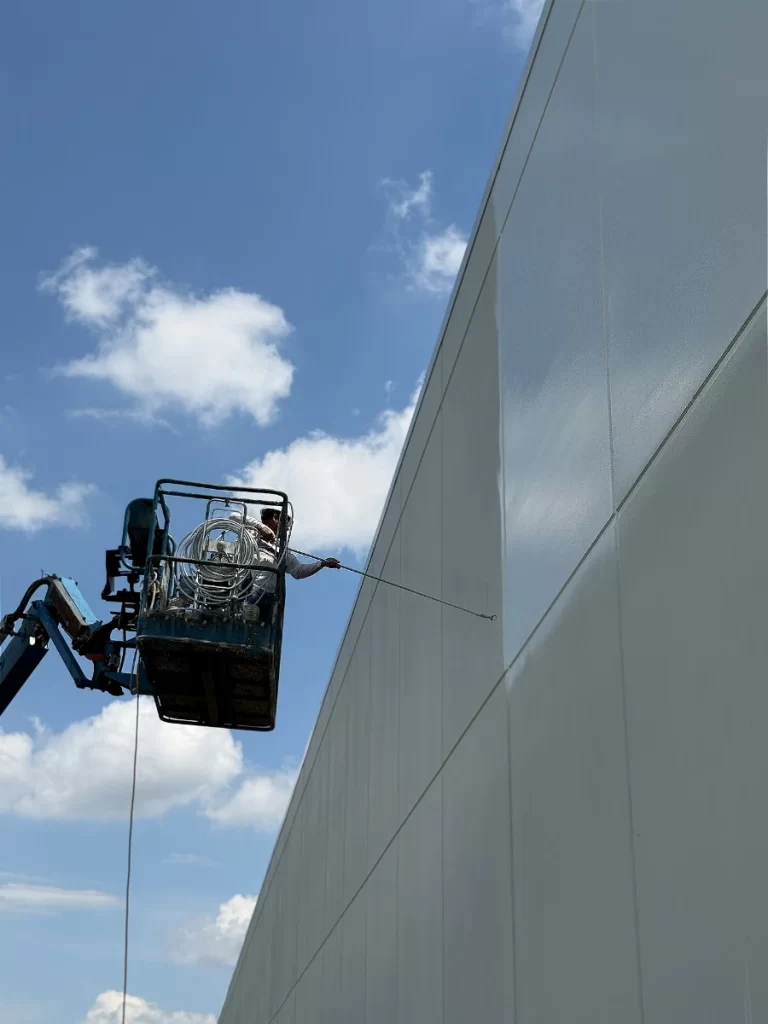 A person on a mechanical lift power washes the exterior wall of a tall building on a sunny day with a blue sky and scattered clouds.