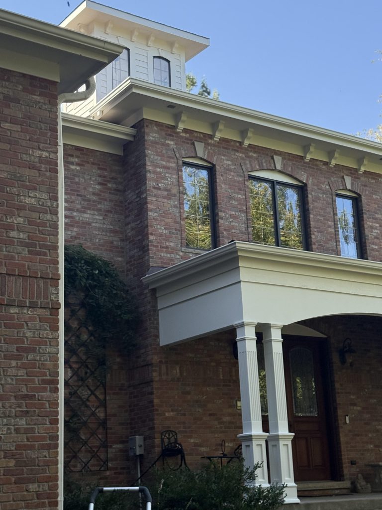A two-story red brick house with black-framed windows, a white cupola, and a covered front entry supported by white columns. Exterior painting highlights the crisp details, while greenery climbs a trellis on the left side.