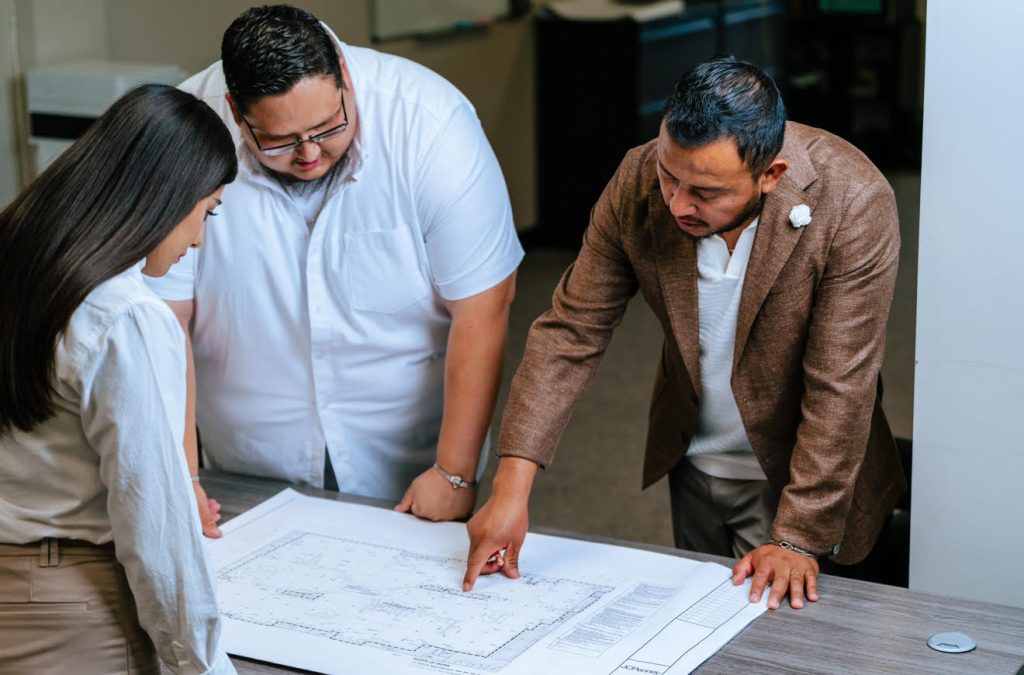 Three people stand around a table, discussing and pointing at a large blueprint labeled "About Metanoia," immersed in planning within an office setting.