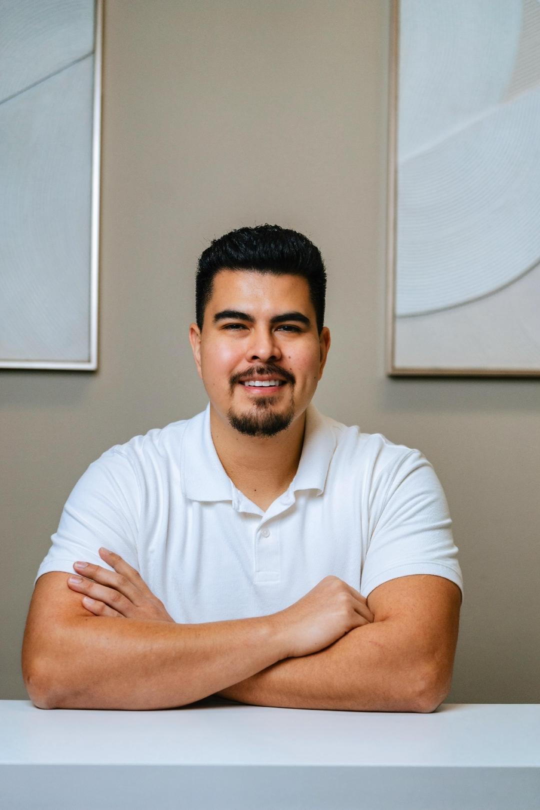 A man with short dark hair and a goatee sits at a white table, smiling with his arms crossed. He is wearing a white polo shirt, and abstract art hangs on a beige wall behind him.